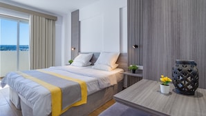 A modern hotel room with a large bed, a bedside table with a lamp, a small table with a vase of flowers, and a view of the city through the window. - Toxotis Hotel  (Paralimni)