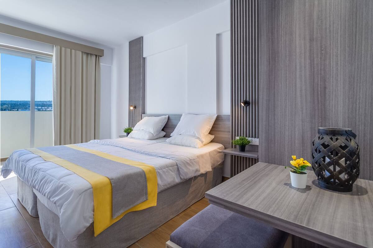 A modern hotel room with a large bed, a bedside table with a lamp, a small table with a vase of flowers, and a view of the city through the window.