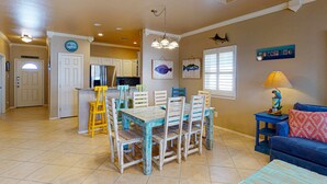 A dining area with a wooden table and chairs, a kitchen in the background, and a blue sofa with a lamp. - Shared Pool, Near Beach, Pet Friendly (Port Aransas)