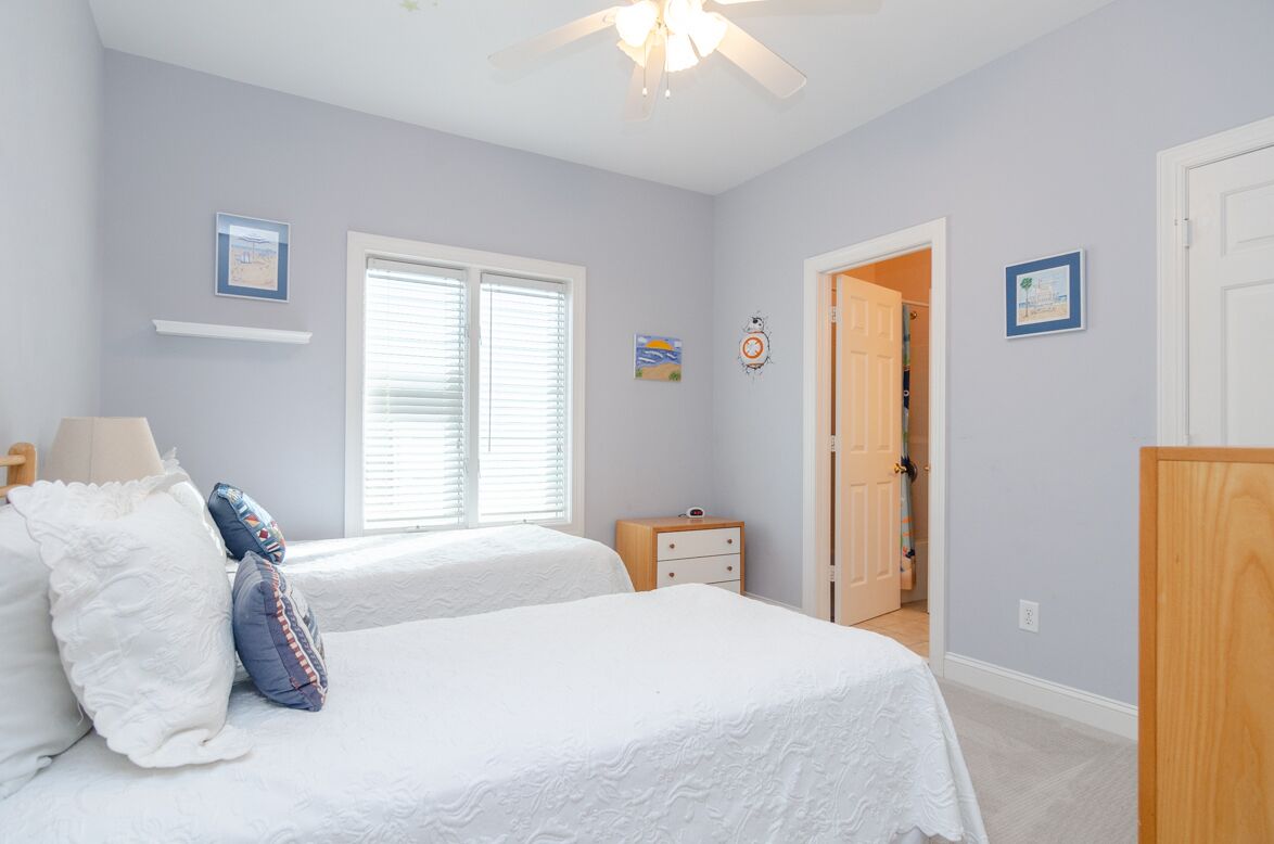 A bedroom with two beds, a ceiling fan, a window with blinds, a nightstand, and a door leading to another room.