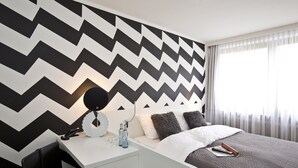 A modern hotel room with a zigzag patterned wall, a white bedside table with a lamp, a remote control, and a bottle of water.