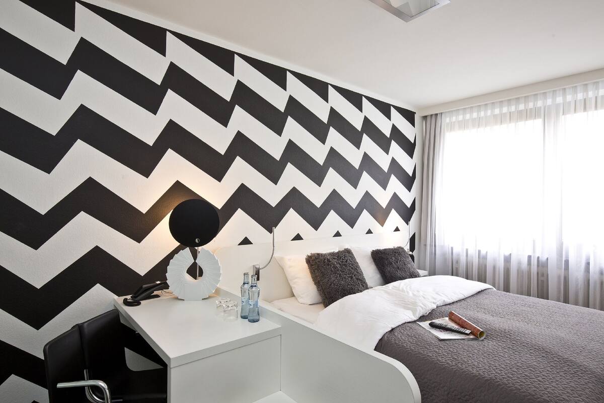 A modern hotel room with a zigzag patterned wall, a white bedside table with a lamp, a remote control, and a bottle of water.