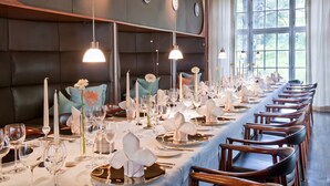 A long dining table set with white tablecloths, silverware, and glassware, with a view of the outdoors through large windows.