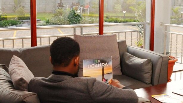 A person sitting on a couch in a room with large windows, reading a book titled "UGANDA". - Pinnacle Hotel Mbarara (Mbarara)