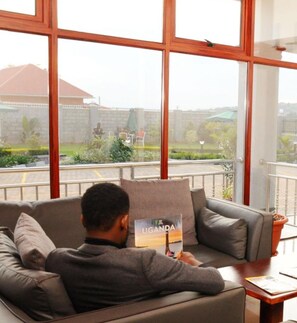 A person sitting on a couch in a room with large windows, reading a book titled "UGANDA". - Pinnacle Hotel Mbarara (Mbarara)