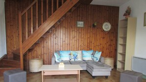 Un salon avec un escalier en bois, un canapé gris orné de coussins en forme d’étoile de mer, une petite table et deux poufs.