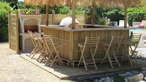 Un espace bar extérieur doté d’un toit de chaume, de chaises en bois et d’un barbecue.