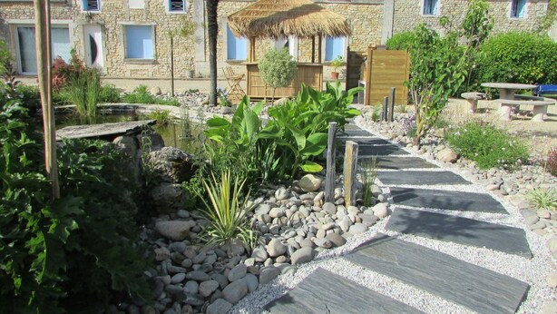 Un bâtiment en pierre doté d’une toiture en chaume, entouré d’un jardin avec un sentier en pierre.