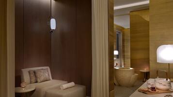 A modern hotel room with a sofa, a small table, a mirror, and a bedside table with a lamp.