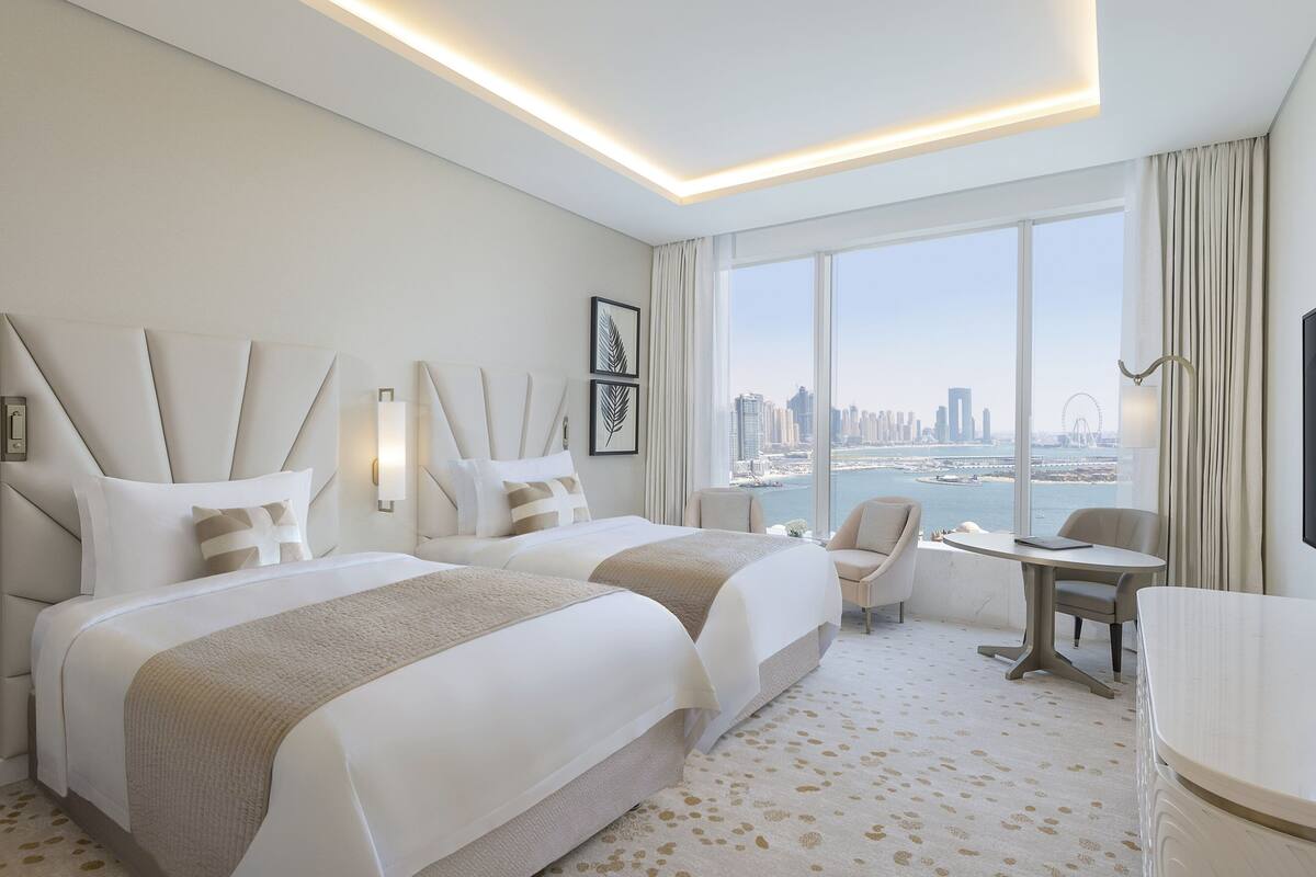 A modern hotel room with two beds, a small table, a chair, and a view of the cityscape and water.
