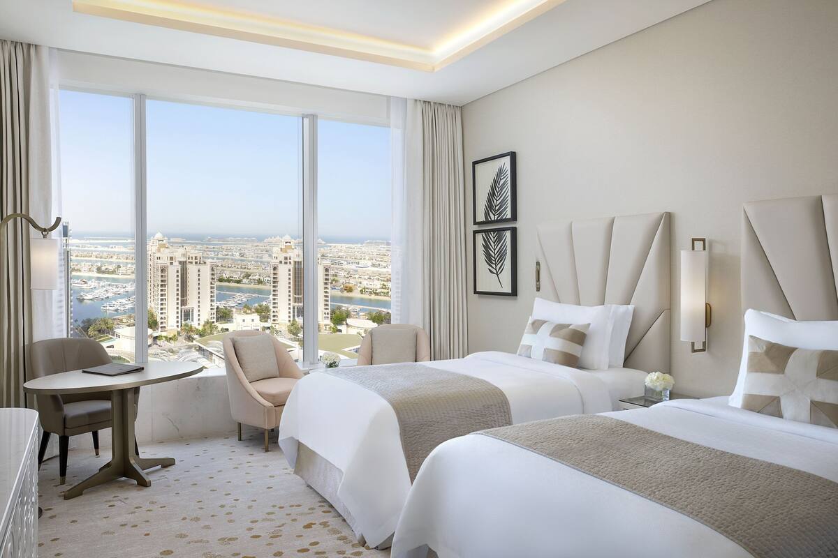 A hotel room with two beds, a small table, a chair, and a view of the city.