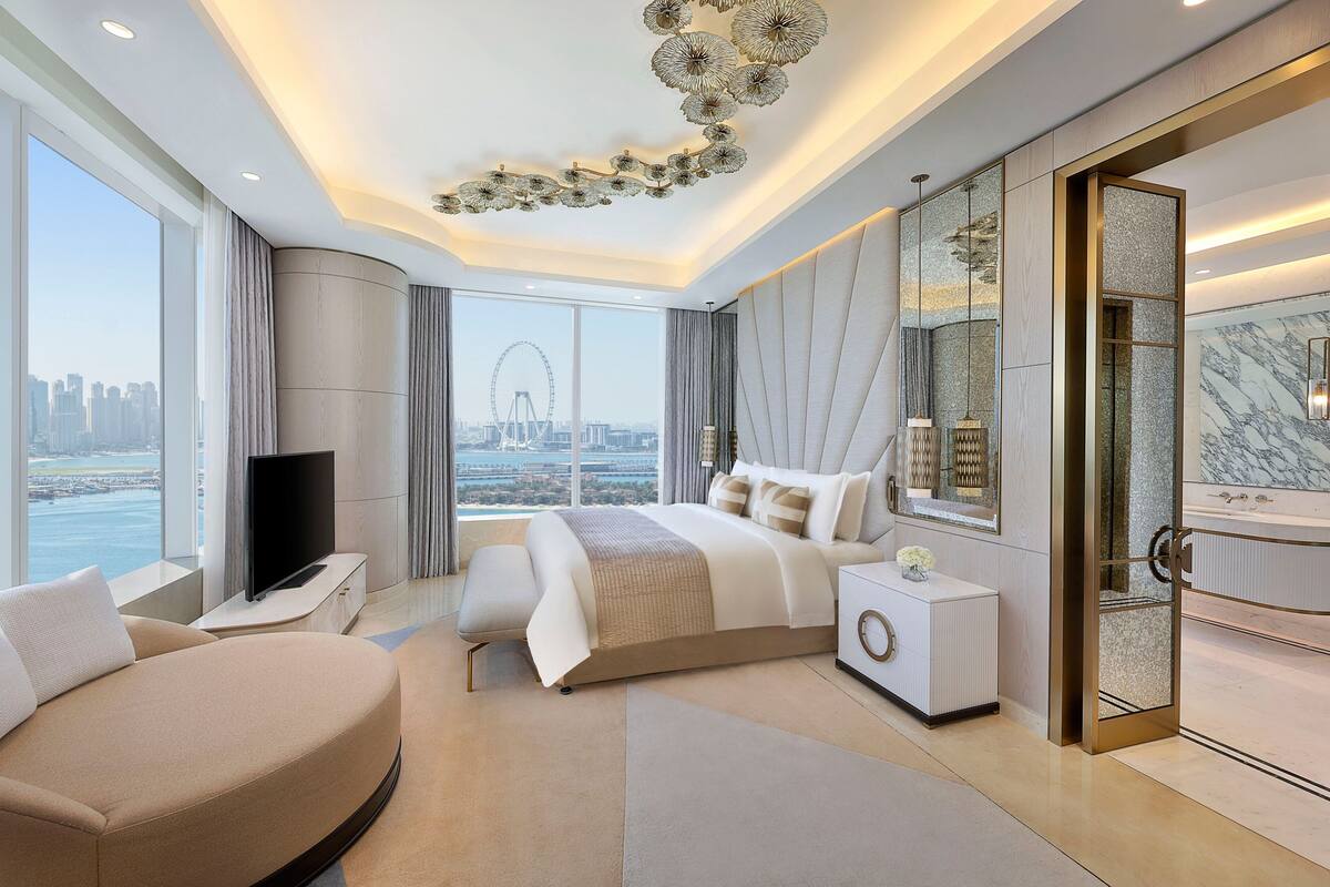 A modern hotel room with a large bed, a TV, a seating area, and a bathroom with a large mirror.