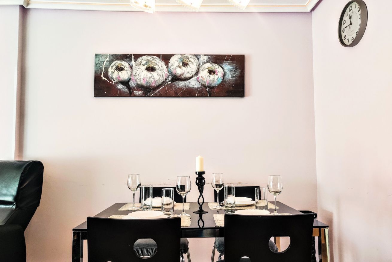 A dining area with a table set for four, a clock on the wall, and a painting above the table.