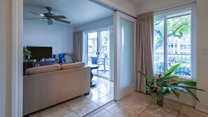 A modern living room with a sofa, a flat-screen TV, and a ceiling fan. There is a sliding glass door leading to a balcony with a view of the neighborhood. - Enjoy A/C, walk to beach/dining/shopping, from the Heart of Kapa'a! (Kapaa)