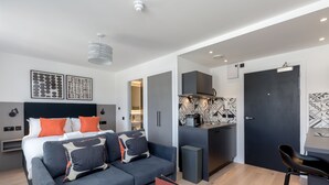 A modern hotel room with a grey sofa, a bed with orange and white bedding, a kitchenette, and a dark door. - Destiny Scotland Market St Apartments (Edinburgh)