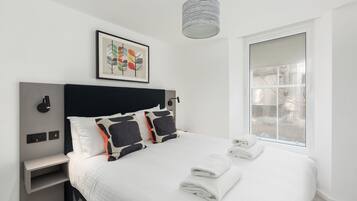 A modern hotel room with a large bed, a headboard with a black panel, a bedside table with a lamp, a framed artwork above the bed, and a window with a view of buildings.
