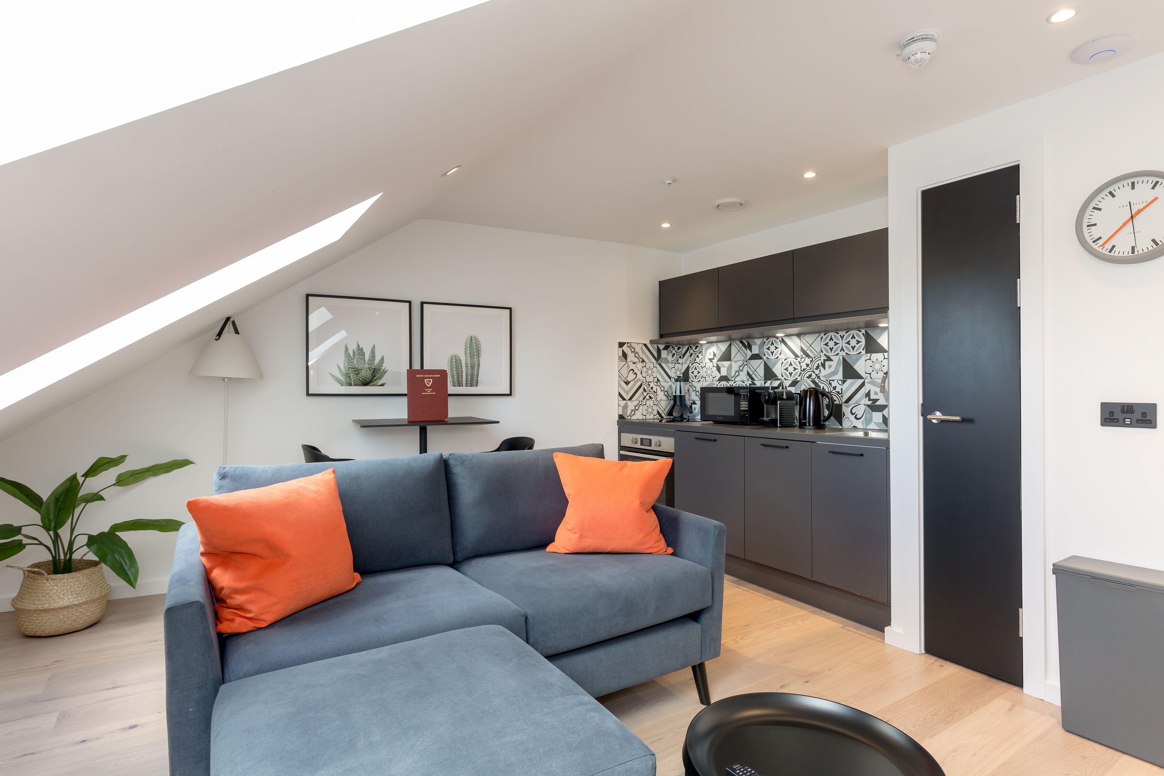 Een moderne woonkamer met een grijze modulaire bank, een oranje kussen, een zwarte fauteuil, een eettafel met stoelen, een keuken met grijze kastjes en een klok aan de muur.