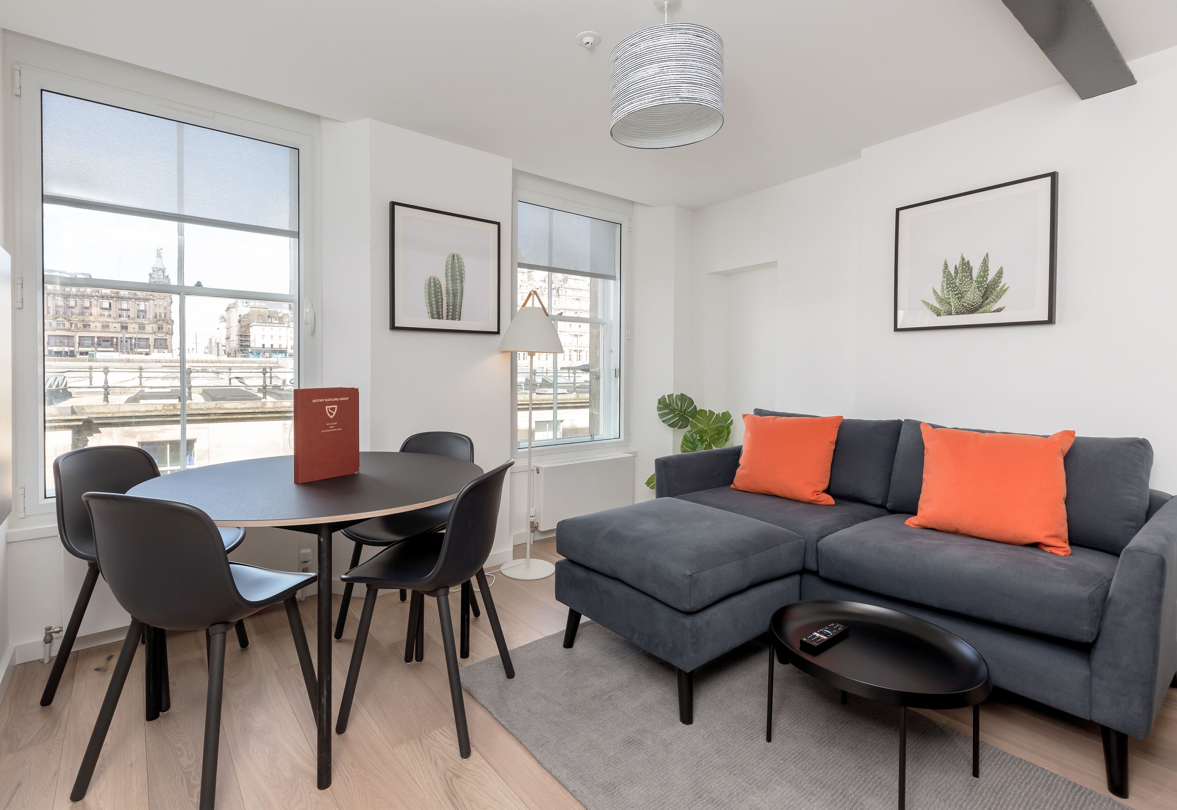 Un salon moderne avec un canapé d’angle gris, des chaises noires, une table ronde et une petite table basse noire. Les murs sont ornés de tableaux encadrés et une grande fenêtre offre une vue sur la ville.