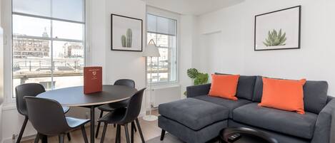 Un salon moderne avec un canapé d’angle gris, des chaises noires, une table ronde et une petite table basse noire. Les murs sont ornés de tableaux encadrés et une grande fenêtre offre une vue sur la ville.