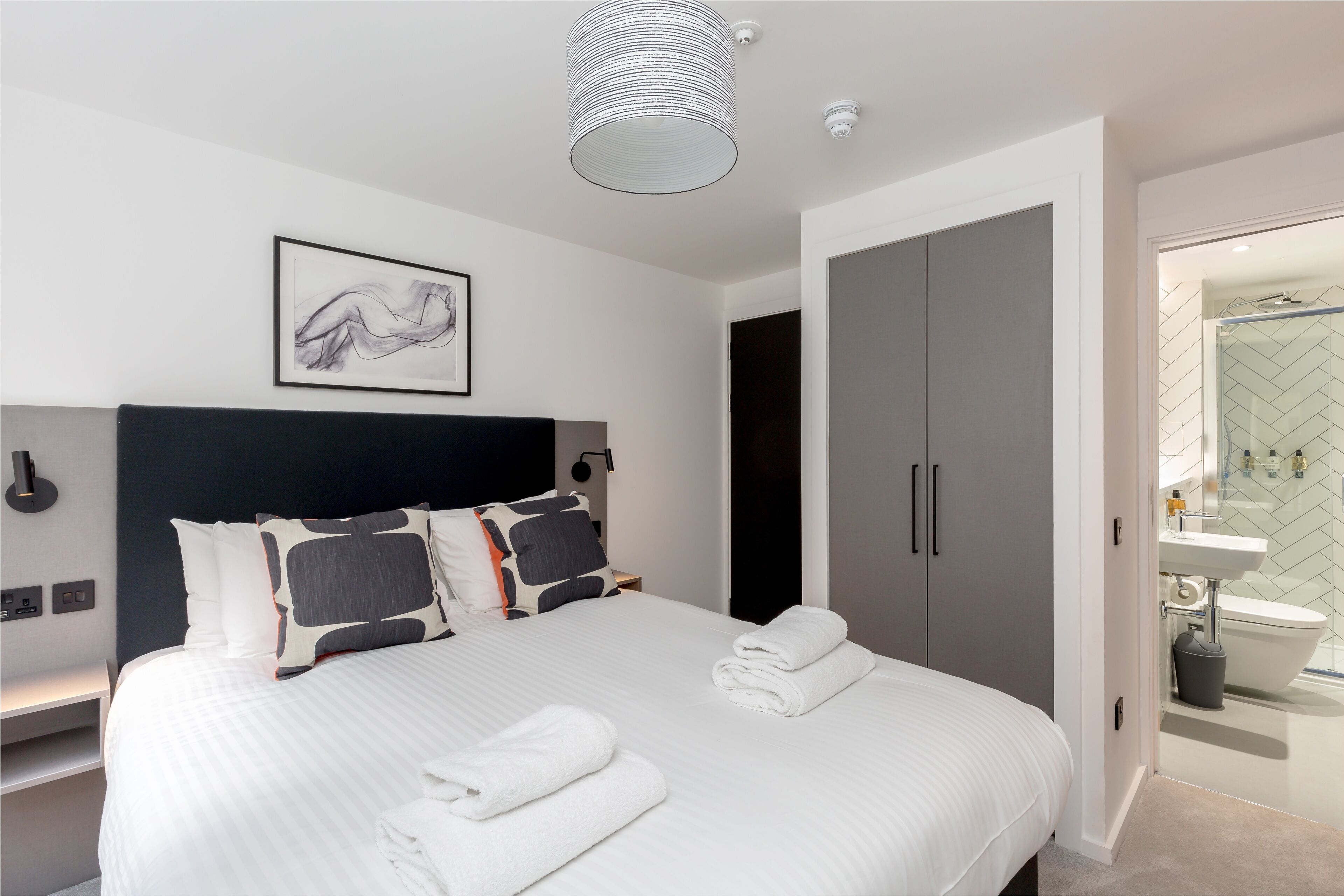 Una habitación de hotel moderna con una cama grande, cabecero, una obra de arte enmarcada, un armario y un baño visible a través de una puerta abierta.