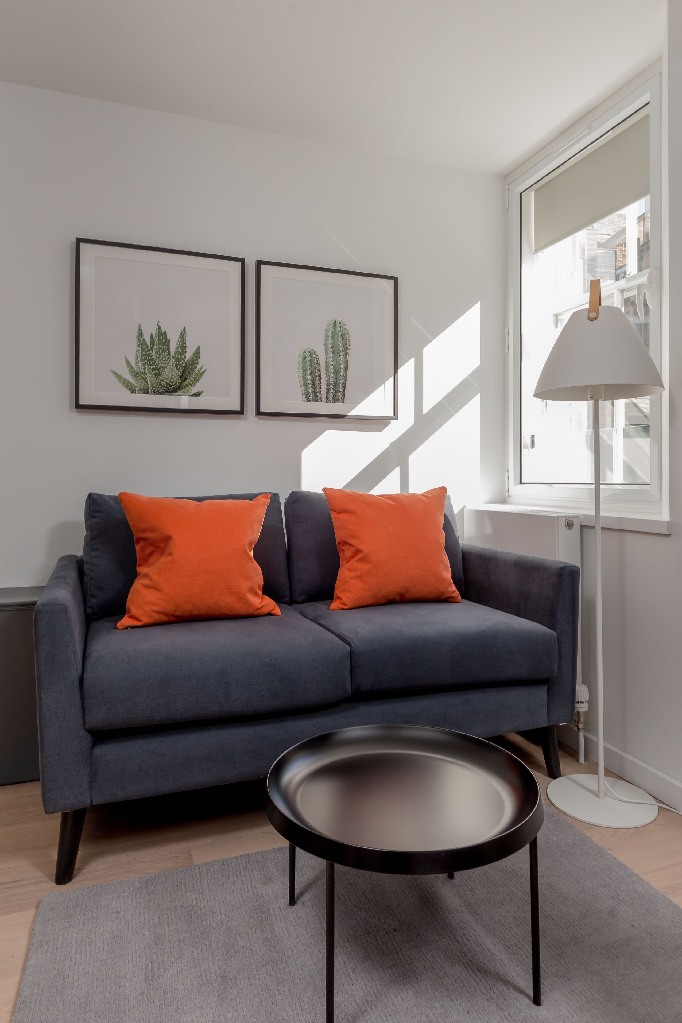 A modern living room with a grey sofa, two orange cushions, a black coffee table, and framed cactus artwork on the wall.