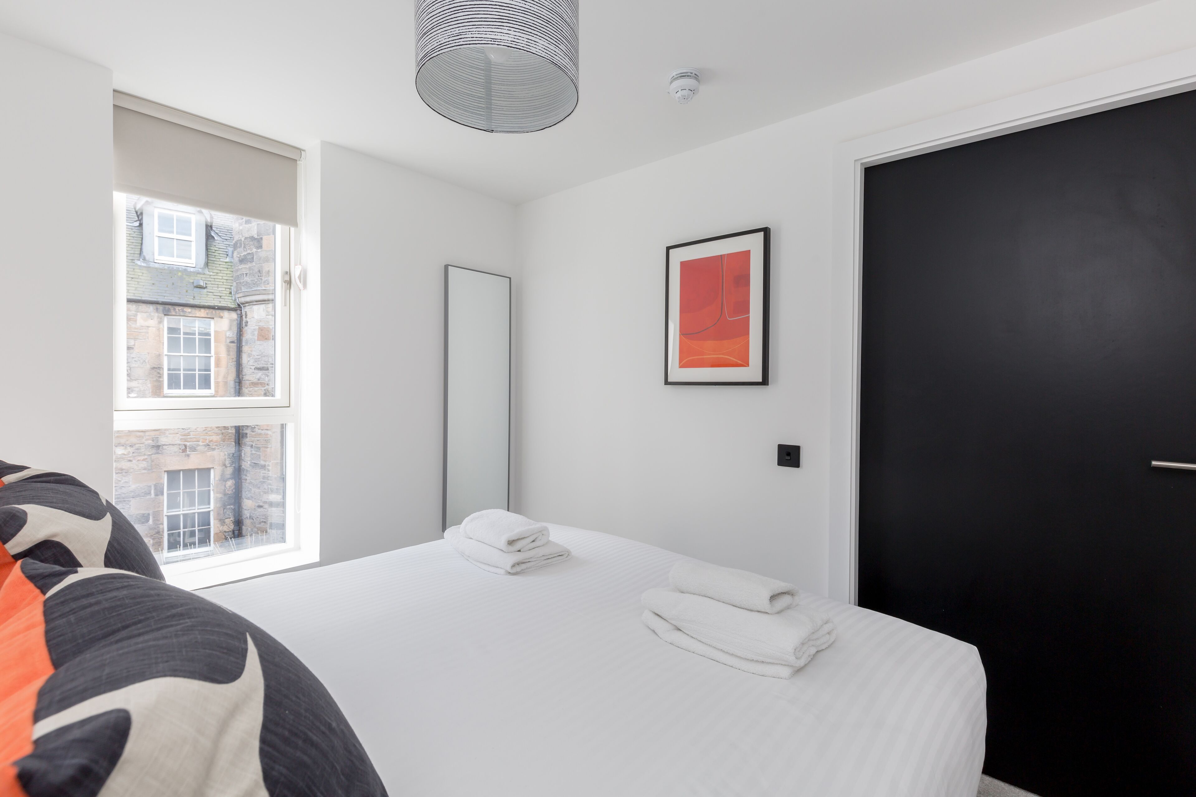 A modern hotel room with a large bed, a black door, a framed artwork, and a window with a view of a building.