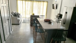 A modern kitchen with a dining area, featuring a long table with bar stools, a refrigerator, and a TV mounted on the wall. - The Best of Home - Tranquil, Comfort & Convenient (Beausejour)