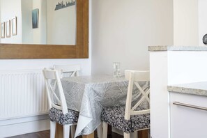 Un tavolo da pranzo con tovaglia a motivi floreali, sedie bianche con cuscini neri a fantasia floreale, uno specchio appeso al muro e un mobiletto bianco con maniglia in metallo.