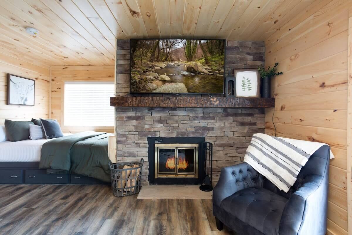 A cozy cabin interior with a fireplace, a bed, a sofa, and a television mounted above the fireplace.