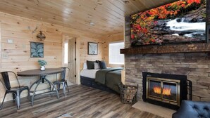 Ein gemütliches Blockhaus-Interieur mit Kamin aus Naturstein, einem Esstisch mit Stühlen, einer Couch und einem großen, an der Wand befestigten Fernseher, der eine Wasserfallszene zeigt.
