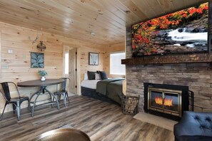 A cozy cabin interior with a stone fireplace, a dining table with chairs, a sofa, and a large wall-mounted TV displaying a waterfall scene. - Extraordinary Mountain Views/Indoor Pool & Hot Tub (Gatlinburg)