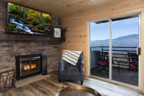A cozy room with a fireplace, a comfortable armchair, and a large window overlooking a deck with outdoor furniture. - Extraordinary Mountain Views/Indoor Pool & Hot Tub (Gatlinburg)