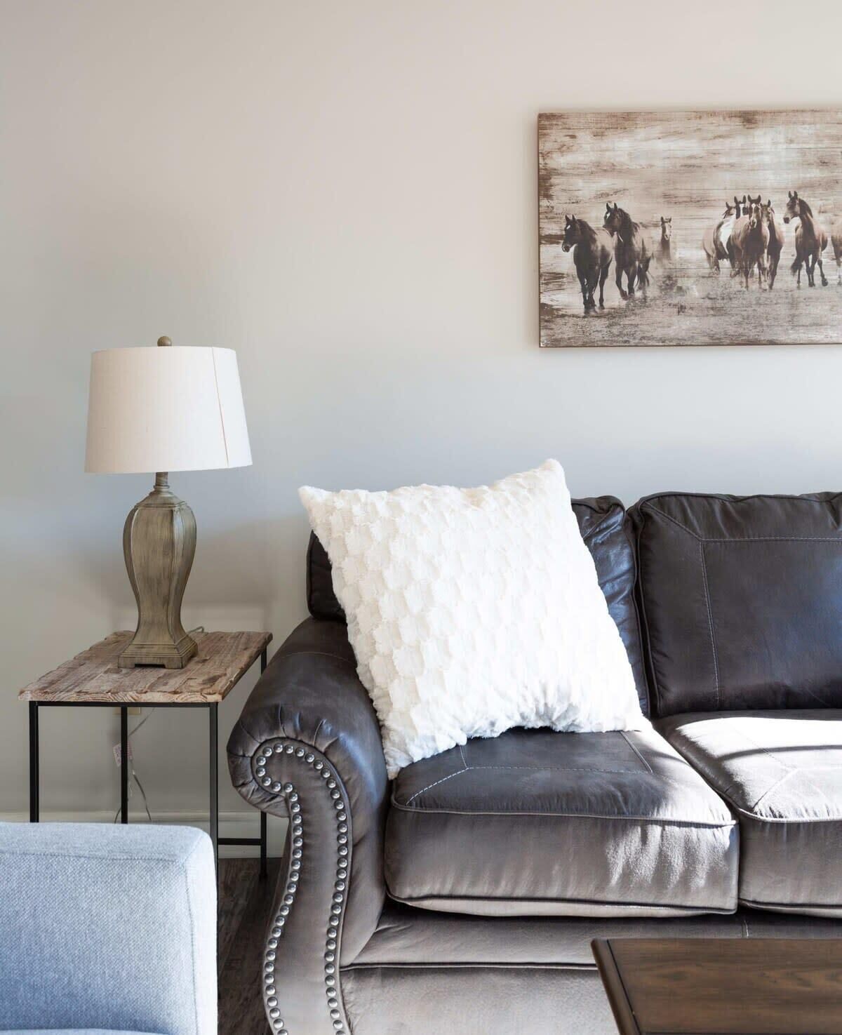 Een woonkamer met een leren bank, een witte kussen, een lamp op een houten tafel en een aan de muur hangend schilderij van paarden.