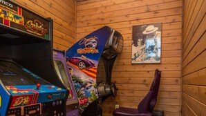 Une petite pièce aux murs en bois et au sol carrelé, qui contient une vieille borne d’arcade et une console de jeux violette.