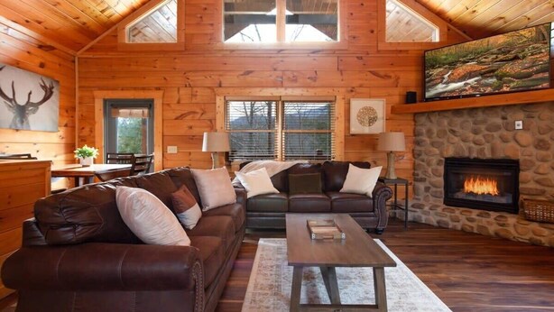 L’intérieur d’une cabane en rondins, avec une cheminée en pierre, des canapés en cuir et un grand téléviseur fixé au mur.