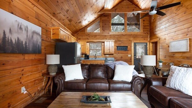 Un acogedor interior de cabaña con techo de madera, una zona de comedor con mesa y sillas, un sofá de cuero marrón y una cocina.