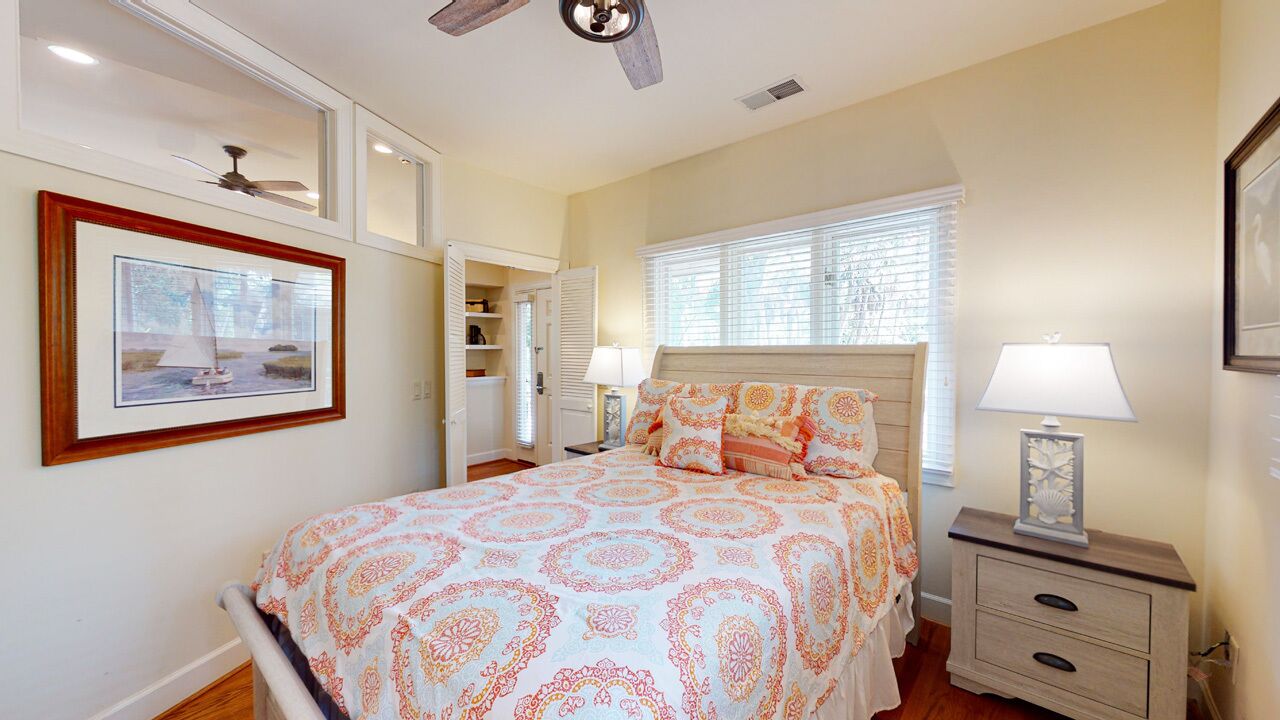 A bedroom with a bed, bedside tables, a ceiling fan, a window with blinds, and a framed picture on the wall.