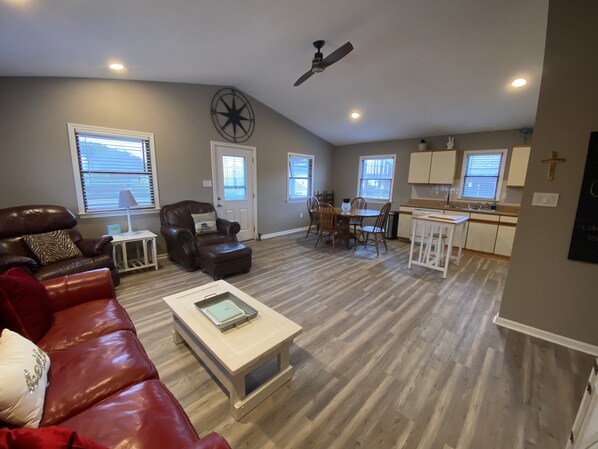 A spacious living room with a leather sofa, a coffee table, and a dining area with chairs and a table. There is a kitchen area with cabinets and a window. - The Sound Castle in KDH (Kill Devil Hills)