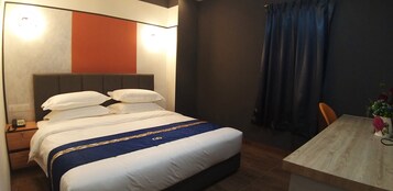 A hotel room with a large bed, a bedside table with a lamp, a small table with a vase of flowers, and a window with curtains.