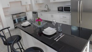 A modern kitchen with white cabinets, a stainless steel refrigerator, a microwave, an oven, and a dining area with a black countertop, bar stools, and a glass vase with flowers. - The Best of Home - Tranquil, Comfort & Convenient (Beausejour)