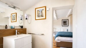 A modern bathroom with a white sink, wooden vanity, and a mirror. There is a door leading to a bedroom with a bed and a framed picture on the wall. - SUITE SAINTE VICTOIRE (Aix-en-Provence)