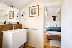 A modern bathroom with a white sink, wooden vanity, and a mirror. There is a door leading to a bedroom with a bed and a framed picture on the wall. - SUITE SAINTE VICTOIRE (Aix-en-Provence)
