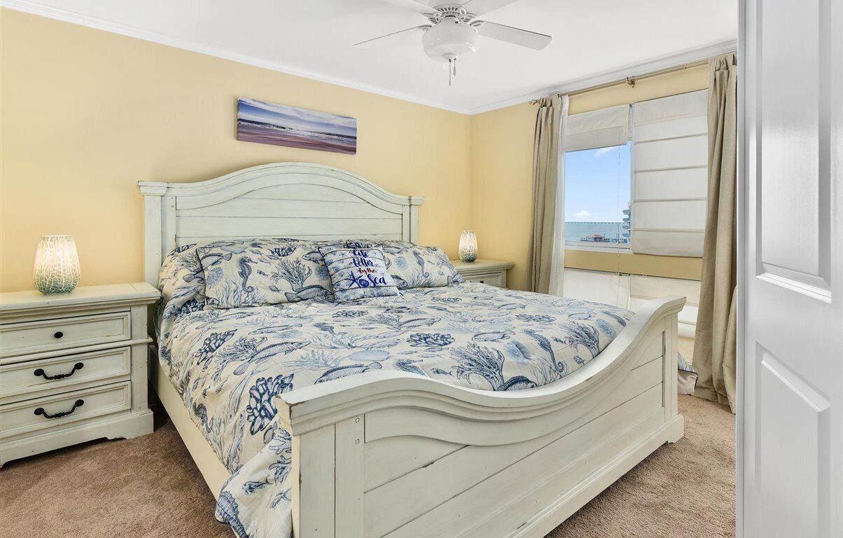 A bedroom with a bed, nightstands, a ceiling fan, a window with curtains, and a painting on the wall.