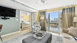 A spacious living room with a grey sofa, a patterned armchair, a flat-screen TV, and a balcony with a view of buildings. - 2 BR 2 BA Unit with Beautiful Bay Views (Ocean City)