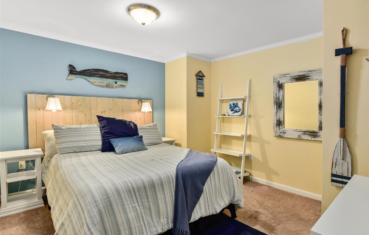 Une chambre à coucher avec une couvre-lit à rayures, une tête de lit en bois, une table de chevet, une étagère avec des objets décoratifs et un miroir fixé au mur.