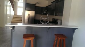 Une cuisine avec des meubles de couleur sombre, un plan de travail blanc et deux tabourets en bois.