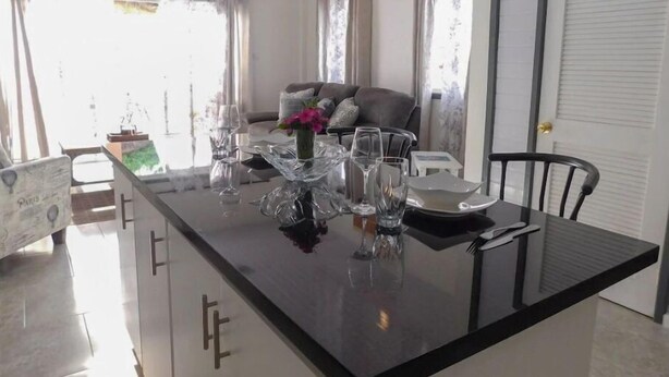 A modern kitchen with a black countertop, white cabinets, and a dining area with a table set for two. - The Best of Home - Tranquil, Comfort & Convenient (Beausejour)