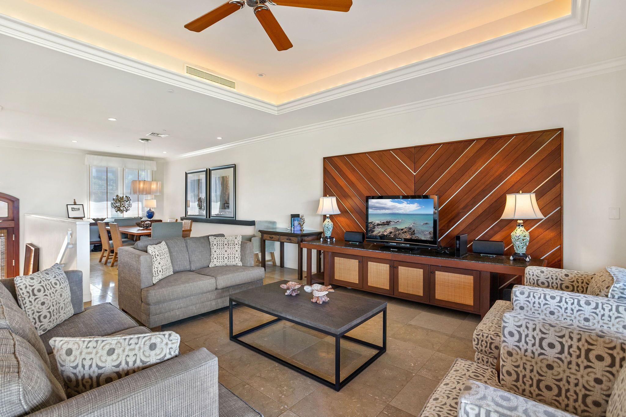 A spacious living room with a modern design, featuring a large flat-screen TV, a wooden accent wall, and a ceiling fan.