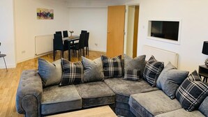 Una sala de estar con un sofá seccional, una mesa de comedor con sillas, un televisor montado en la pared y una puerta que da a otra habitación.