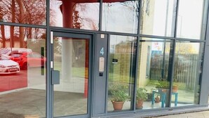 Entrada de un edificio con puertas de vidrio, una placa con un número y plantas en macetas afuera.